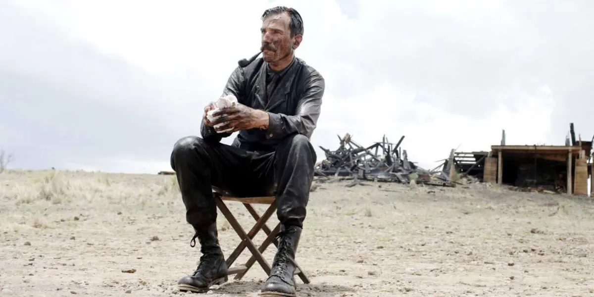 Daniel sitting on a chair outside covered in oil and smoking a pipe.