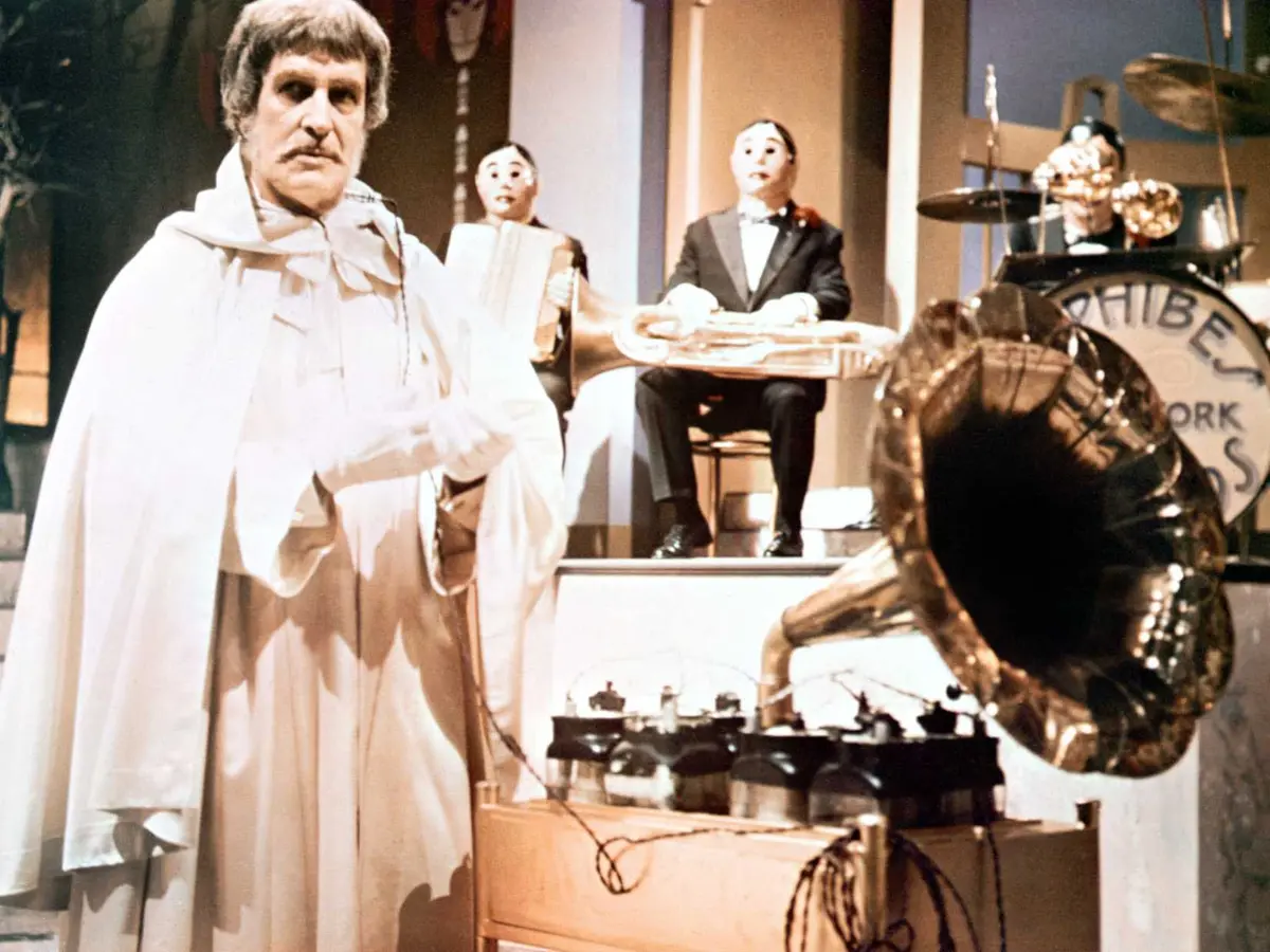 Vincent Price in a white cape stands next to a gramophone