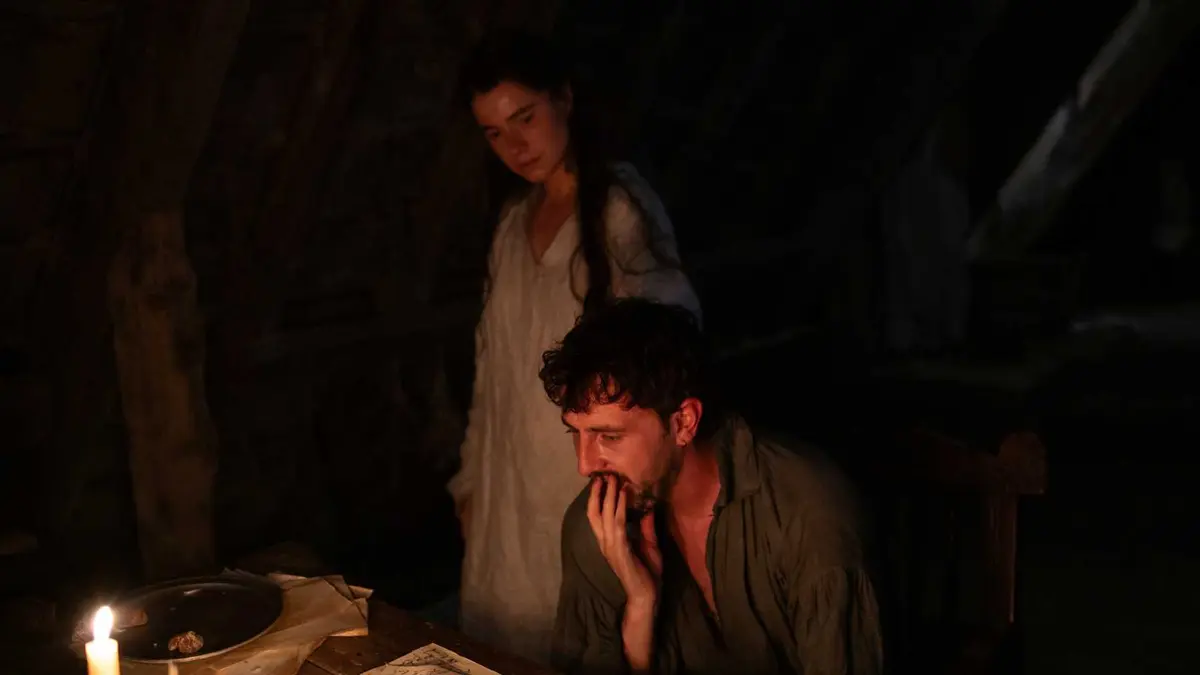 Agnes looks over Wills shoulder while he writes in their darkened befroom via candle light in Hamnet