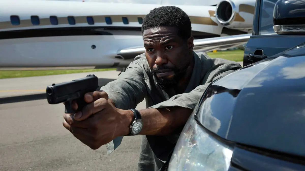 Yahya Abdul-Mateen II's Creasy aiming a gun while hiding behind a truck in front of a jet in Man on Fire