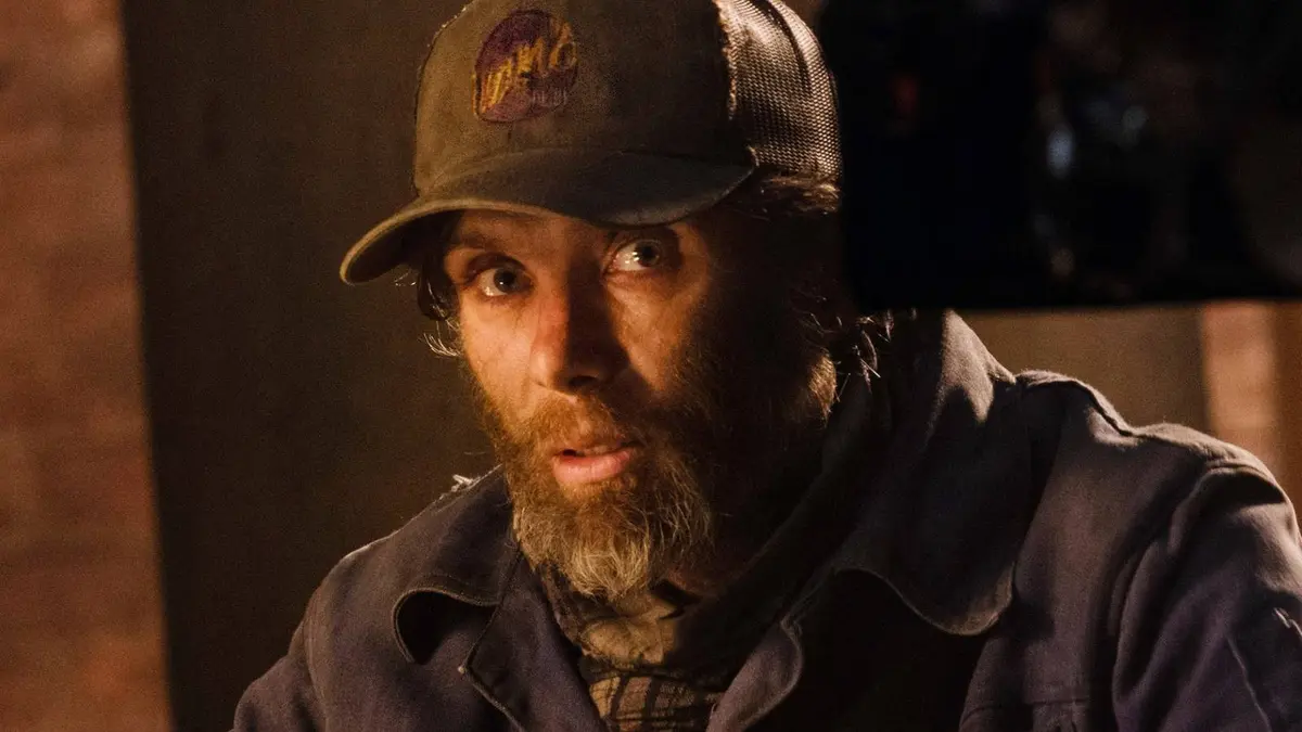 A bearded Cillian Murphy wearing a ballcap and sitting in a dark room