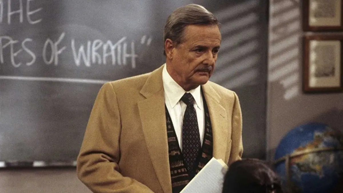 An older male teacher stands in front of a blackboard that reads The Grapes of Wrath