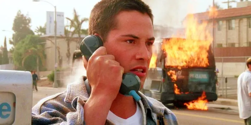 Keanu Reeves as Jack Traven talking on a payphone while a vehicle is on fire in the background