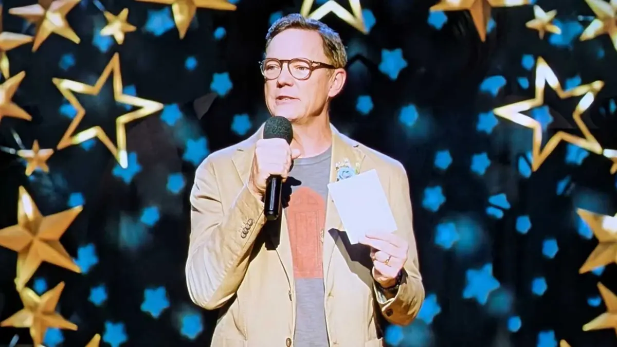 Matthew Lillard as Principal Henry Grayle wearing a tan blazer, black t-shirt with a red door, holding a microphone and an envelope on a stage